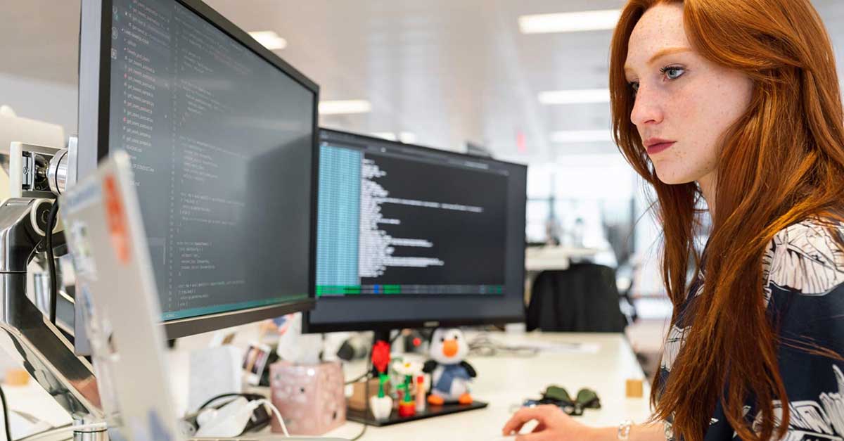 Redhaired woman sitting at desk looking at code and logs on 2 monitors