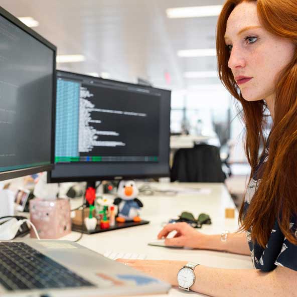 red haired woman working on code at desk 2 monitors FintechOS